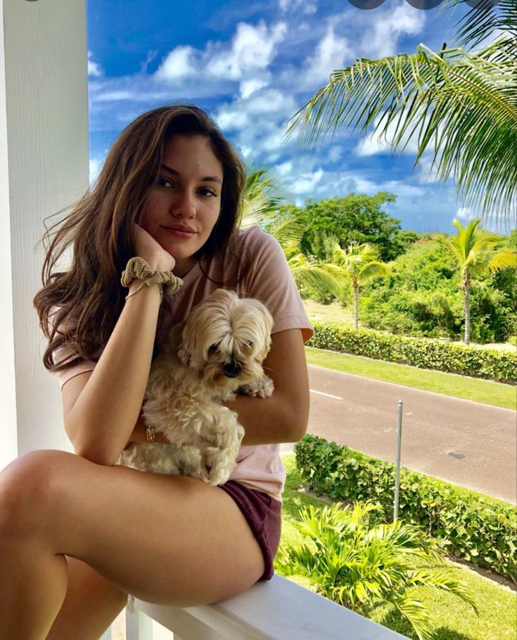 a woman sitting on a porch holding a small dog and looking at the camera with a blue sky in the background