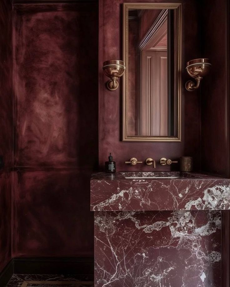 a bathroom with red walls and marble counter top, gold fixtures on the mirror over the sink