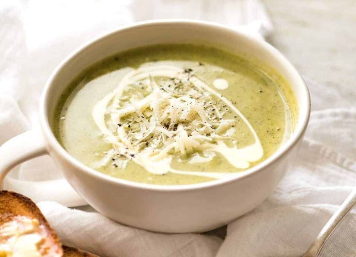 a white bowl filled with broccoli soup next to a piece of bread on a napkin