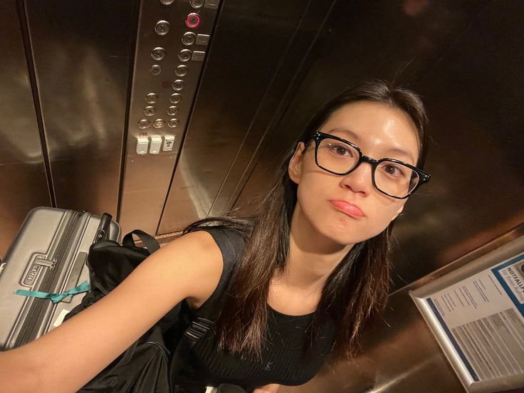 a woman wearing glasses and holding a suitcase in her hand while standing next to an elevator