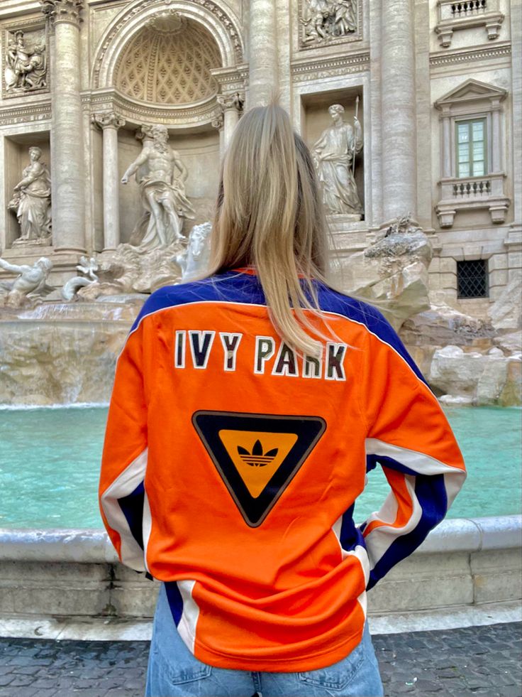 a woman standing in front of a fountain wearing an orange jacket that says ivv park