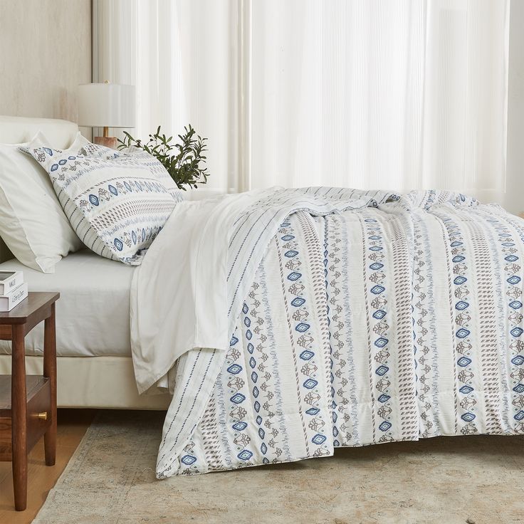 a bed with white sheets and pillows next to a lamp on a table in front of a window