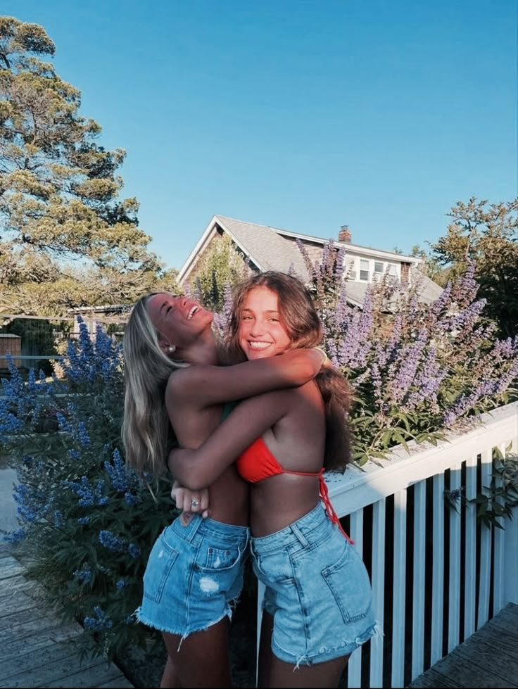 two young women hugging each other in front of purple flowers and a white picket fence