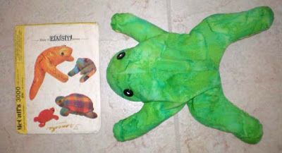 a green stuffed animal laying next to a book on the floor with it's cover pulled up