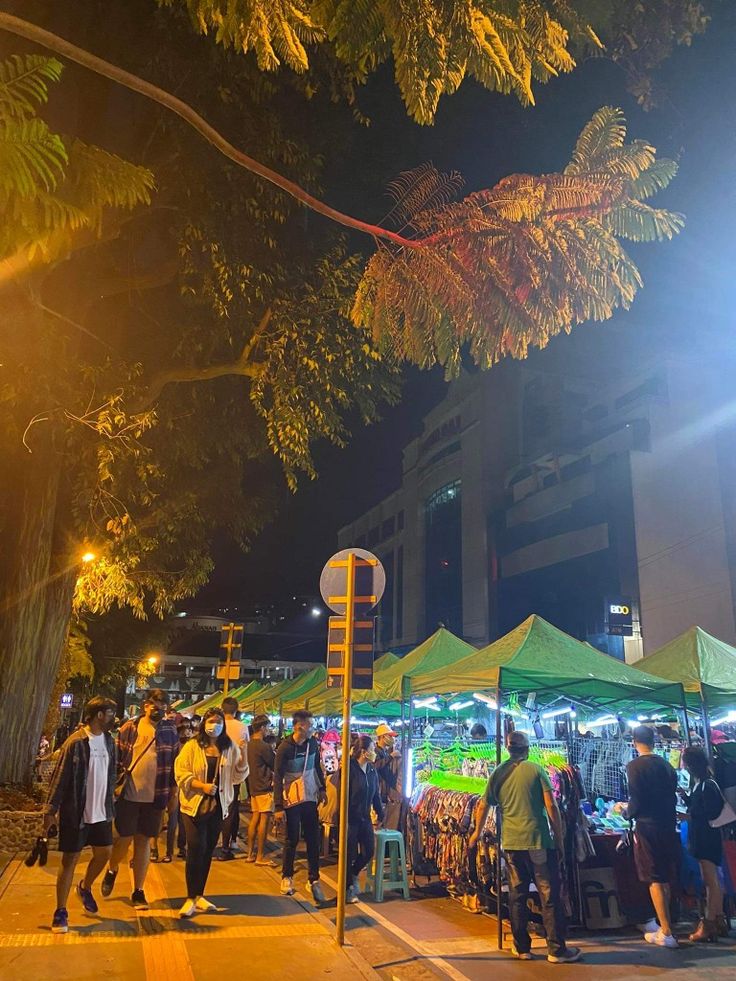 many people are walking around an outdoor market area at night, with tents and lights in the background