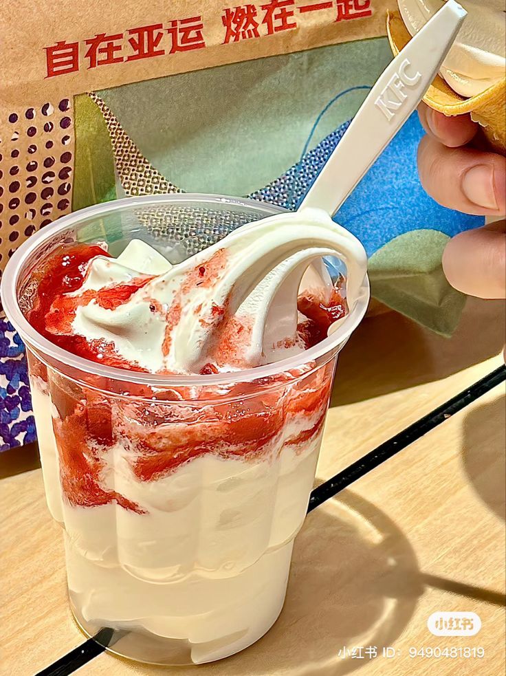 a person holding a spoon in a plastic cup filled with ice cream and strawberries