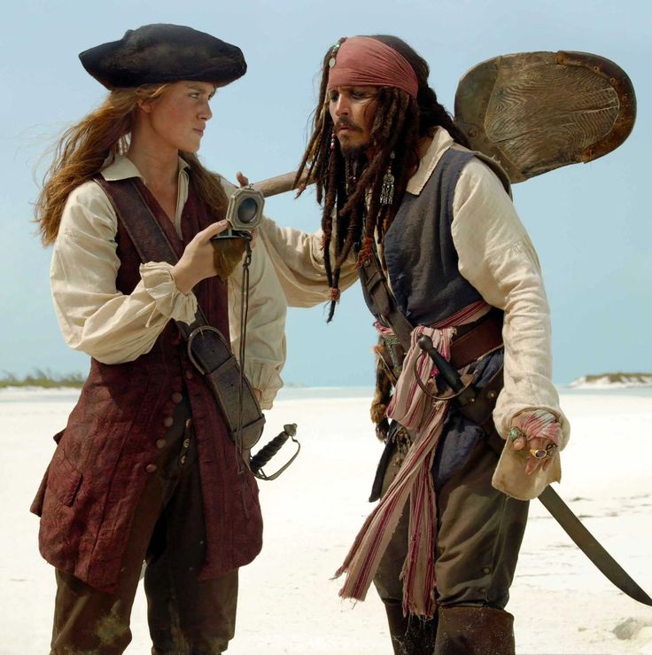 two people dressed in pirate costumes on the beach with one holding a camera and looking at another person's face