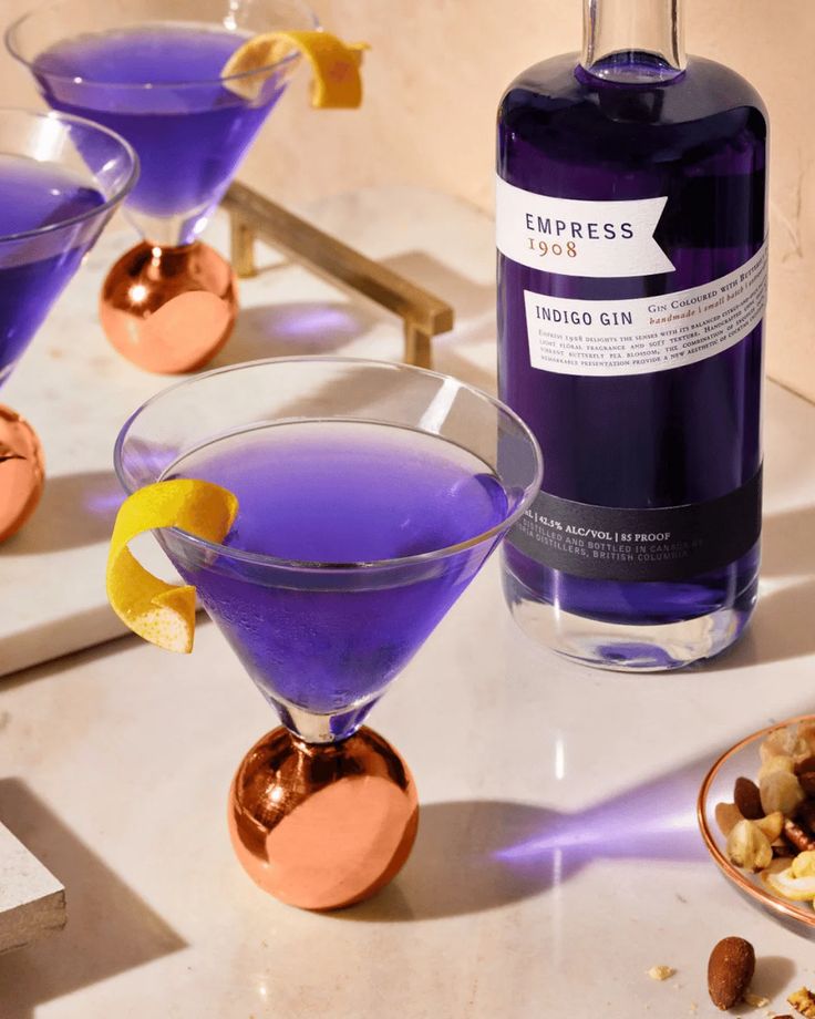 two martini glasses filled with purple liquid next to a bottle of liquor and nuts on the table