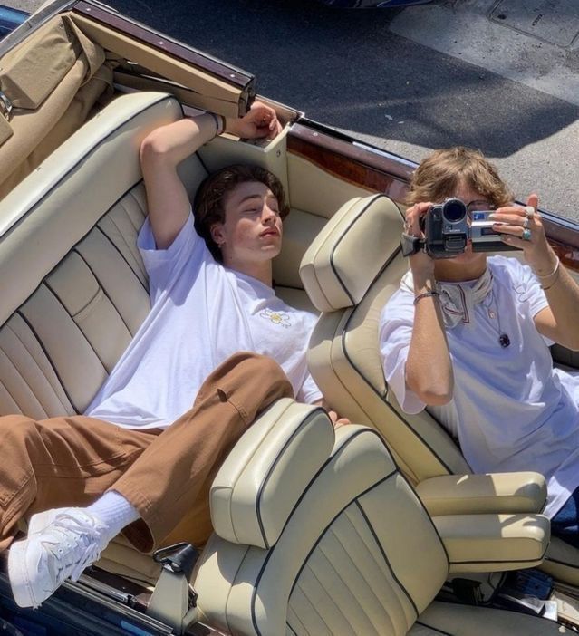 two young men sitting in the back of a convertible car, one holding a camera