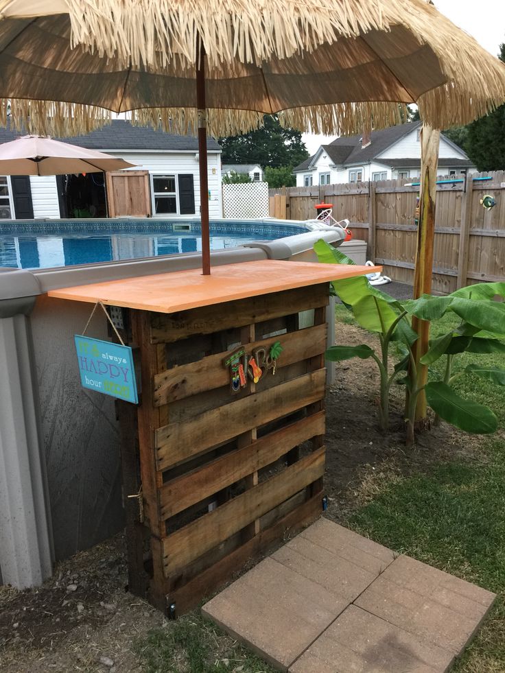 an outdoor bar made out of pallet wood with straw umbrella over it and a pool in the background