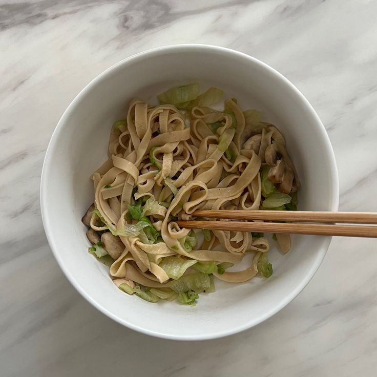 a white bowl filled with noodles and vegetables next to chopsticks on a marble surface