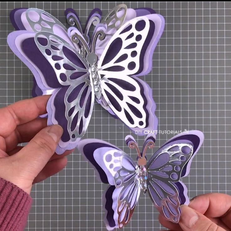 two hands holding purple and white butterflies on top of a gray surface with grids in the background