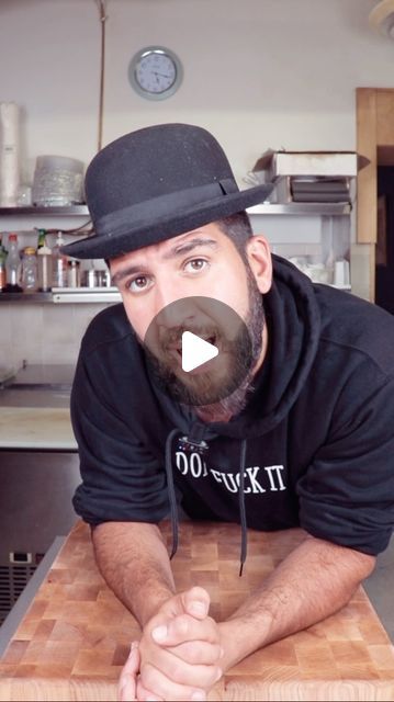 a man with a beard and top hat leaning on a table in a kitchen looking at the camera