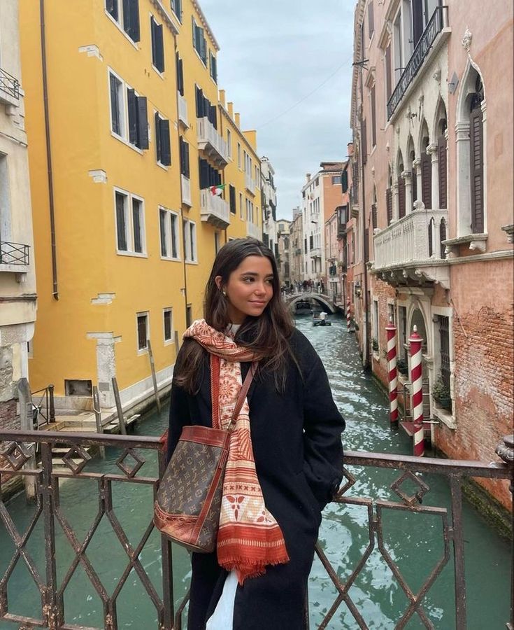 a woman standing on a bridge over a canal with buildings in the background and a gondola passing by