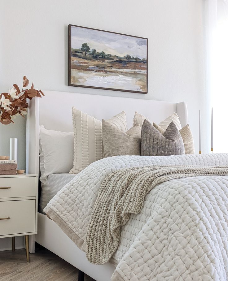 a white bed topped with lots of pillows next to a painting on the wall above it