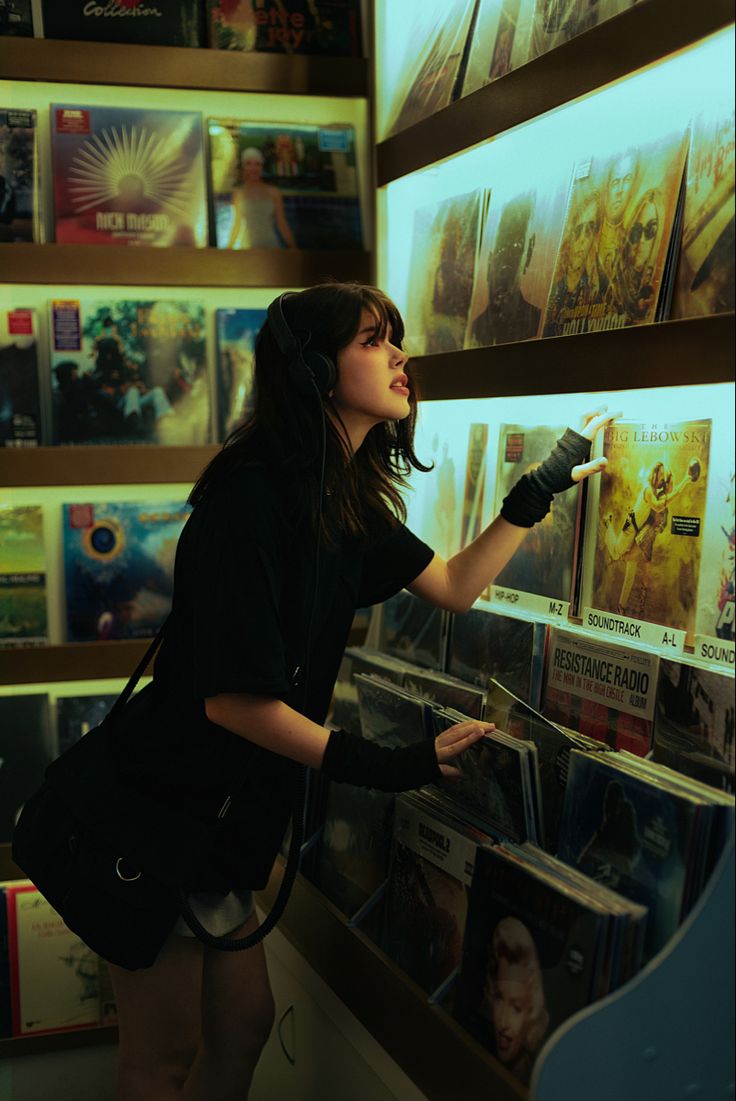 a woman with headphones on looking at records in a record store, she is leaning against the wall