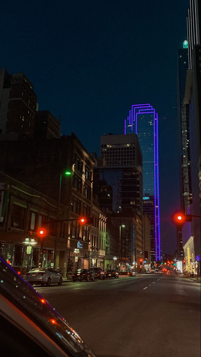 an empty city street at night with cars parked on the side of it and tall buildings in the background