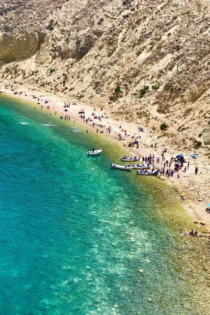 people are standing on the beach next to the water and boats in the blue ocean