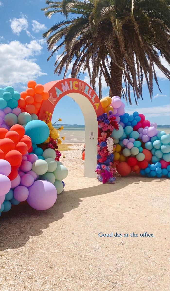 an arch made out of balloons on the beach with a palm tree in the background