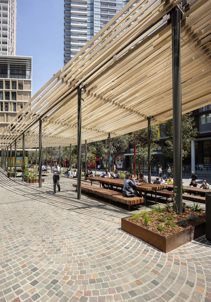 people are sitting on benches under an awning in a public area with tall buildings