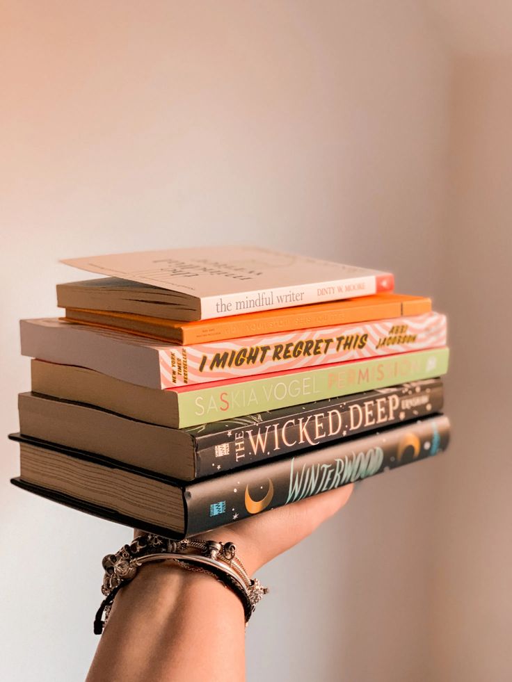 a stack of books held by a woman's hand in front of a white wall