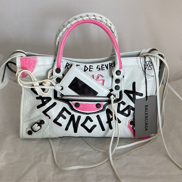 Authentic Balenciaga Limited Edition City S Graffiti Handbag In White With Black And Pink Logo Graffiti. Larger Than The Mini In A Desirable Size Small. Very Roomy And Beautifully Done In A Soft, Supple Leather. Rolled Whip Stitched Double Handles, Front Center Outer Pocket With Attached Framed Mirror. All Dark Bronze Hardware. Detachable And Adjustable Shoulder Strap. Tasseled Zips. Double Inner Slip Pocket And One Zip Pocket. Black Canvas Lining. Eye Catching And Show Stopping In These Bold Co Designer White Shoulder Bag For Spring, Designer Shoulder Bag For Spring, Designer White Top Handle Shoulder Bag, Luxury White Shoulder Bag For Spring, Designer White Bag With Dust Bag, White Designer Bags With Branded Hardware, Balenciaga Small City, Balenciaga Giant City Bag, Balenciaga Mini Bag