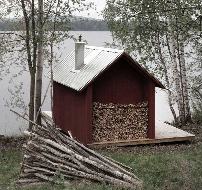 a house made out of firewood next to the water with trees in front of it