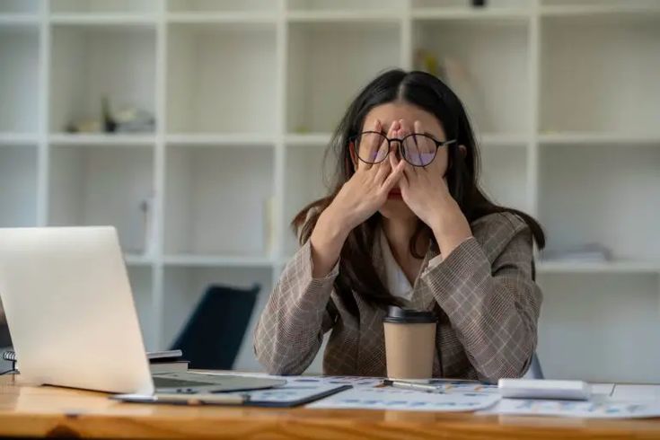 a woman covering her eyes while sitting at a desk with a laptop in front of her