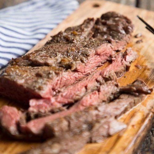 a steak on a wooden cutting board with tongs next to it and a striped dish towel in the background