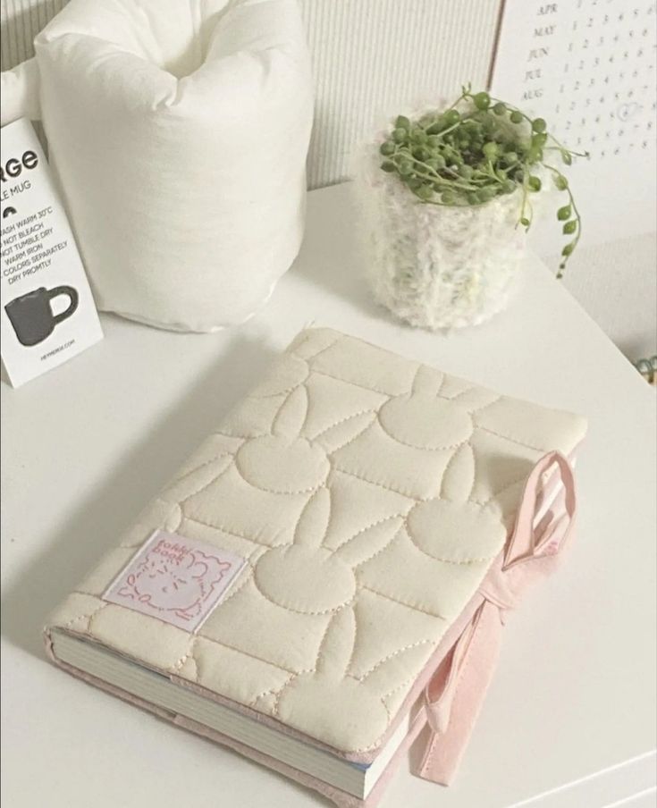 a white table topped with a notebook covered in pink ribbon next to a potted plant