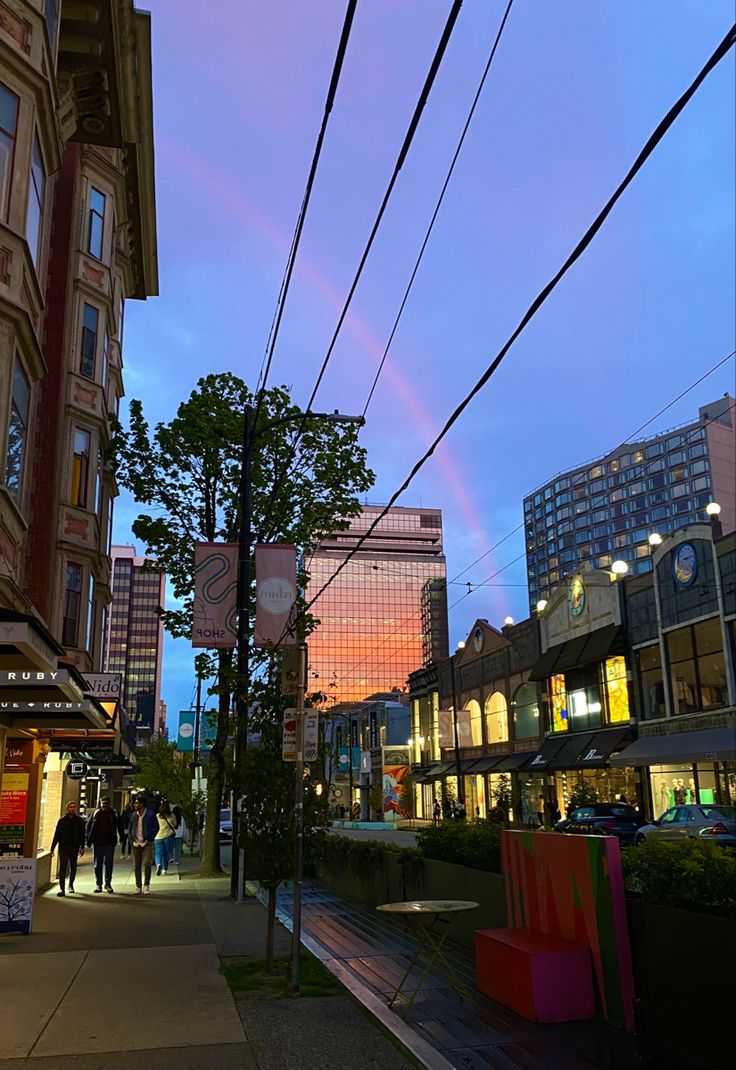 people walking on the sidewalk in front of buildings at dusk with a rainbow in the sky