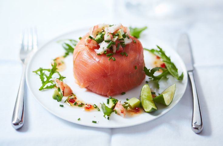 a white plate topped with a piece of salmon covered in toppings next to a fork and knife