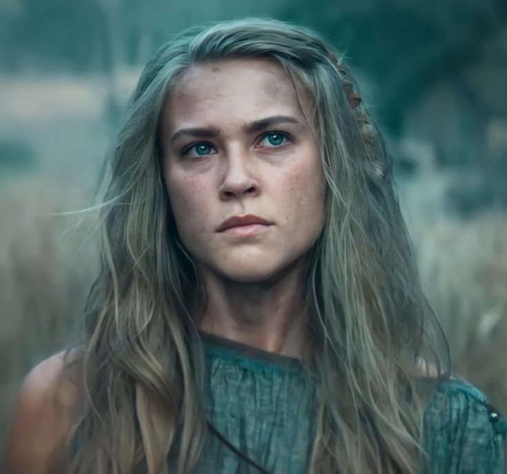 a woman with long blonde hair and blue eyes is staring at the camera while standing in a field