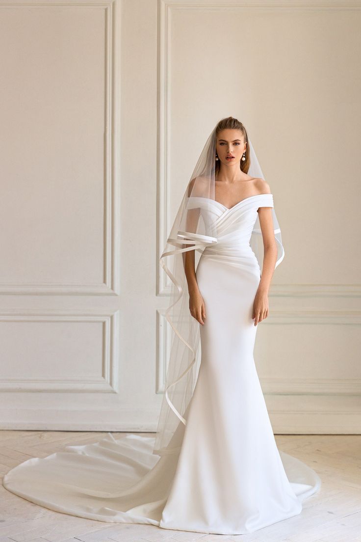 a woman wearing a white wedding dress with a veil on her head and shoulder, standing in front of a wall