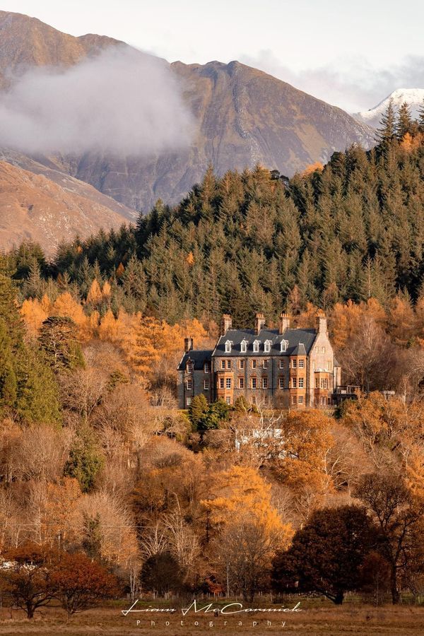 a large house in the middle of a forest with mountains in the background and clouds