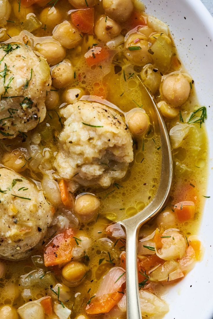 a white bowl filled with meatballs and vegetables next to a spoon on top of a table