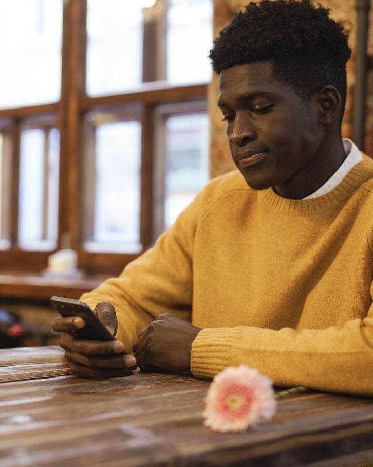 a young man sitting at a table looking at his cell phone while holding a flower