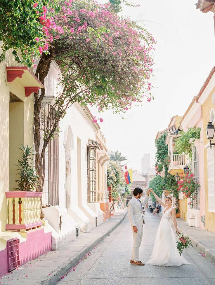 a bride and groom walking down the street in front of colorful buildings with pink flowers