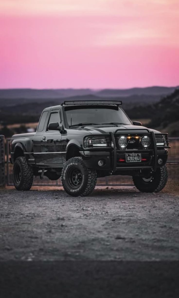 a black truck is parked in front of a fence at dusk with the sun setting