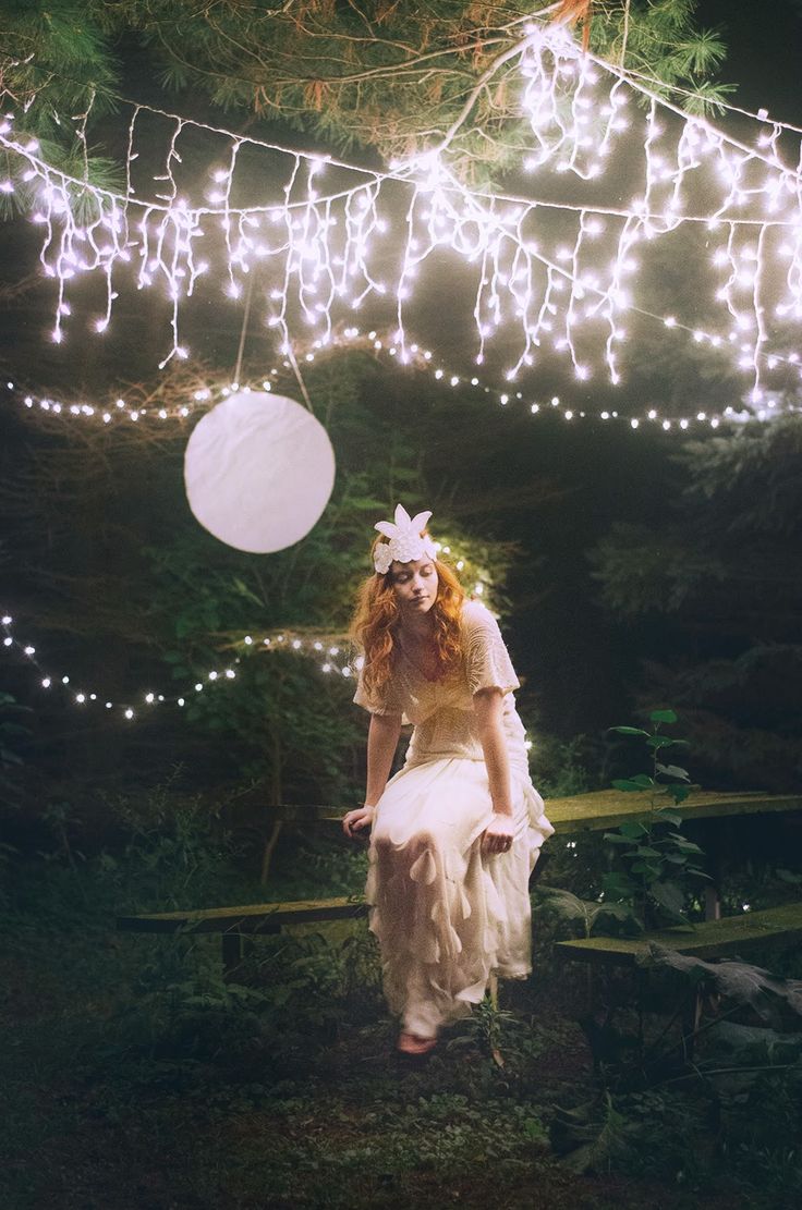 a woman in a white dress is walking through the woods with fairy lights strung above her