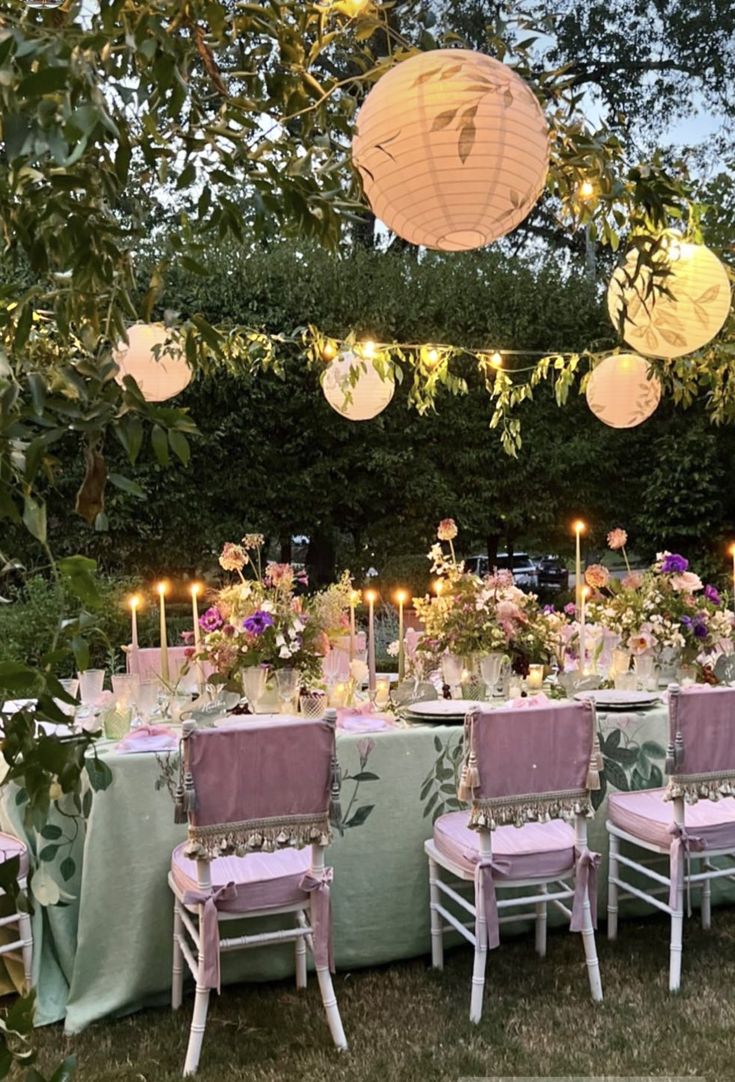 an outdoor dinner table set up with pink chairs and paper lanterns hanging from the trees