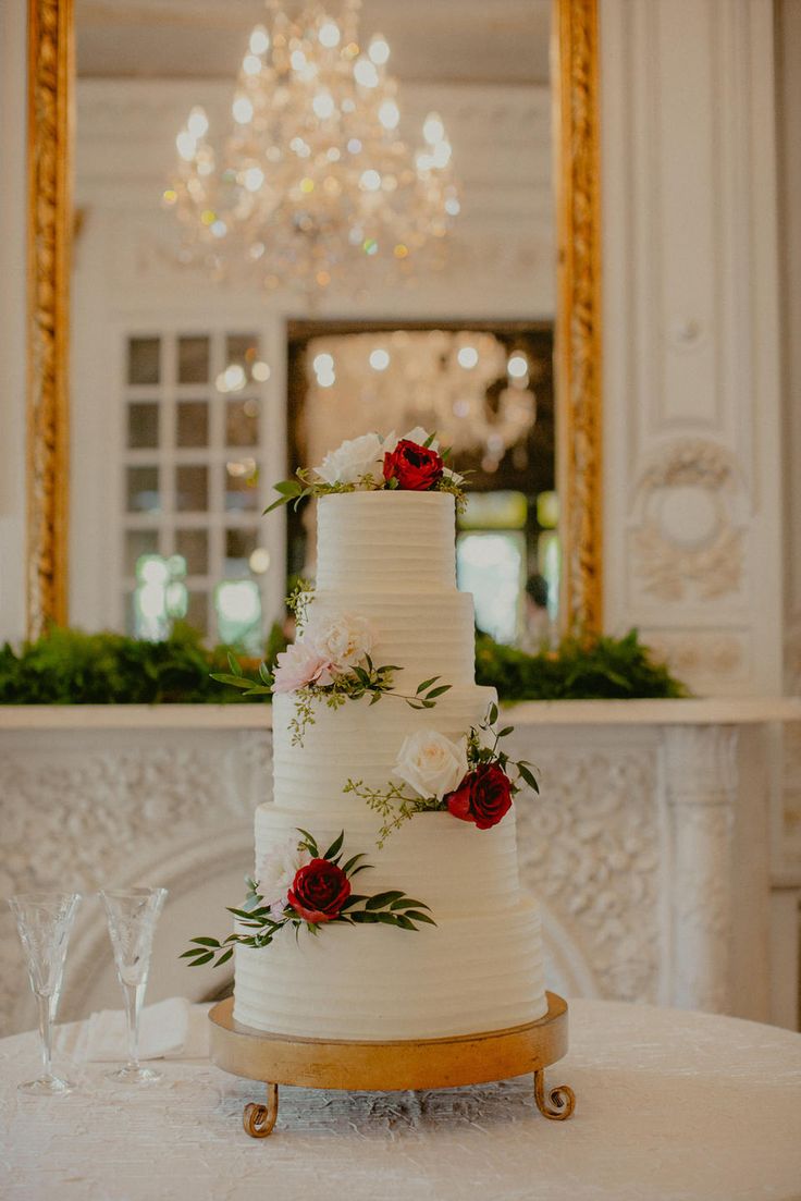 a three tiered wedding cake with red and white flowers on the top is sitting on a table in front of a chandelier