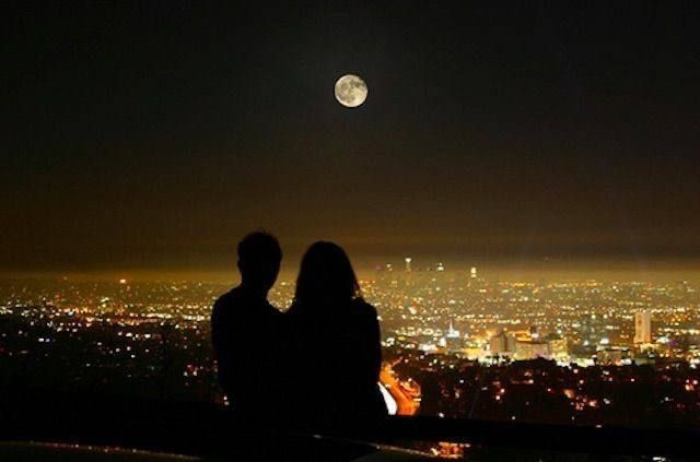 two people are sitting on a ledge looking at the city lights in the night sky