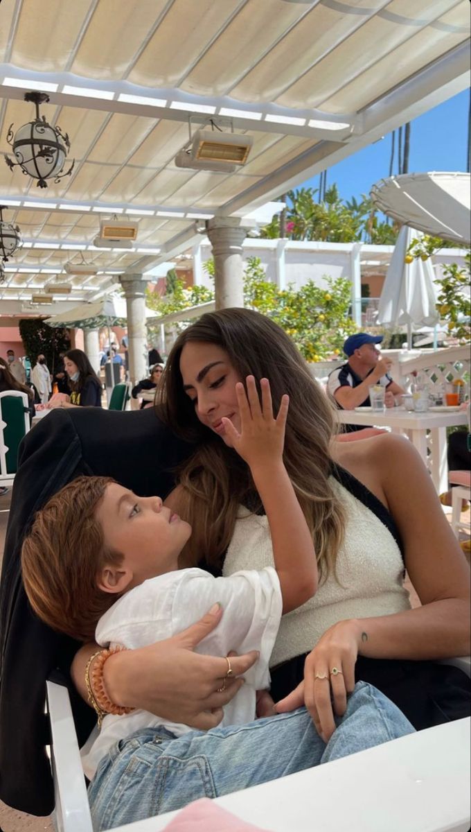 two women and a baby are sitting on a bench in an outdoor cafe, one is holding her hand up to the other's face