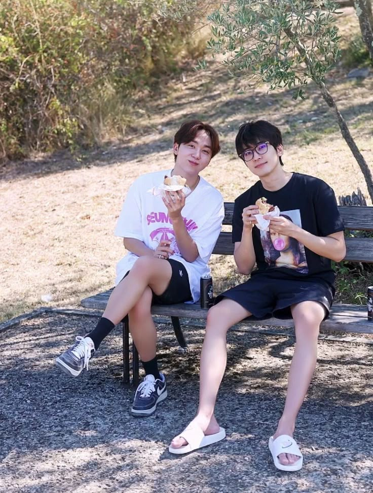 two young men sitting on a bench eating ice cream and drinking soda while one man looks at the camera