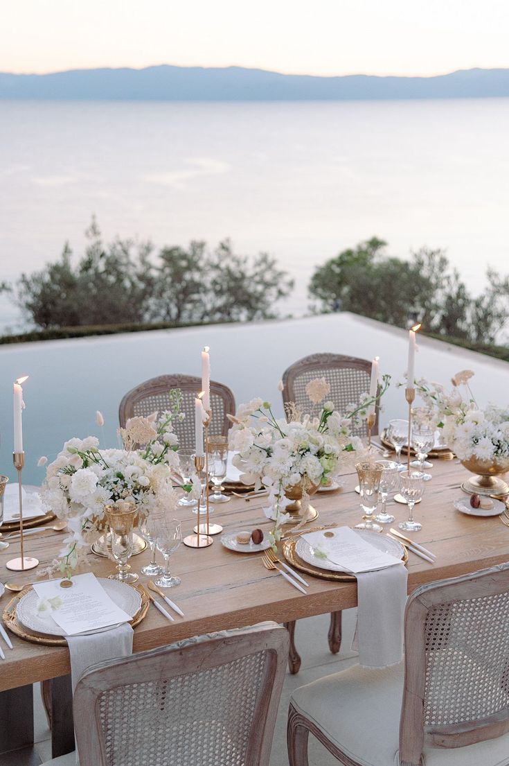 an outdoor table set up with white flowers and candles on it, overlooking the water