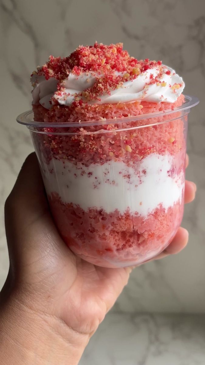 someone is holding up a dessert in a plastic cup with white frosting and red sprinkles