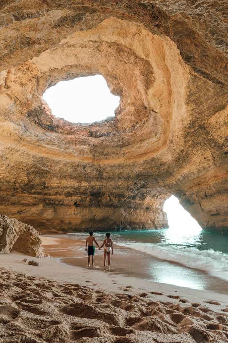 two people holding hands on the beach in front of an arch shaped rock formation with water and sand