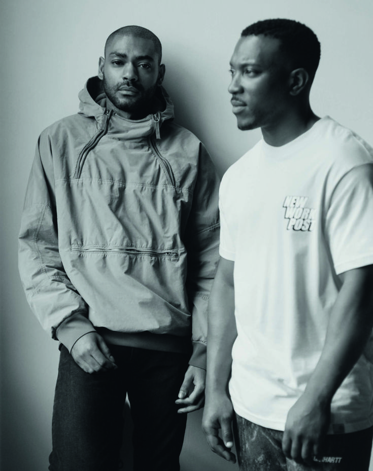 two men standing next to each other in front of a white wall and one is looking at the camera