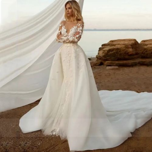 a woman standing on top of a sandy beach wearing a wedding dress with long sleeves